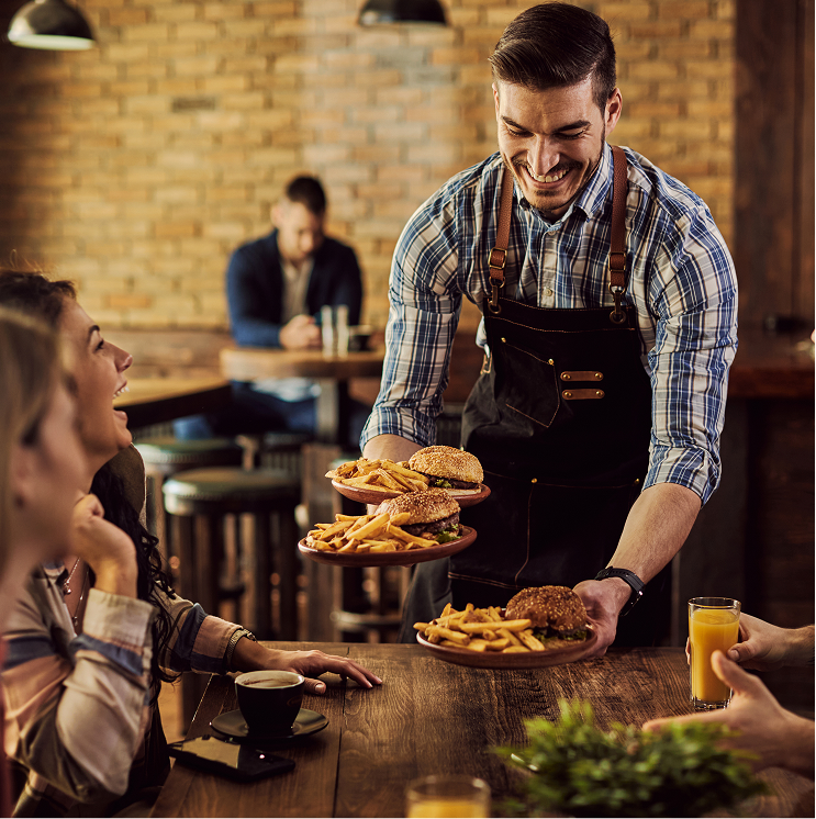 Camarero sonriente trayendo platos con hamburguesas y patatas a la mesa durante una experiencia social POPULIT